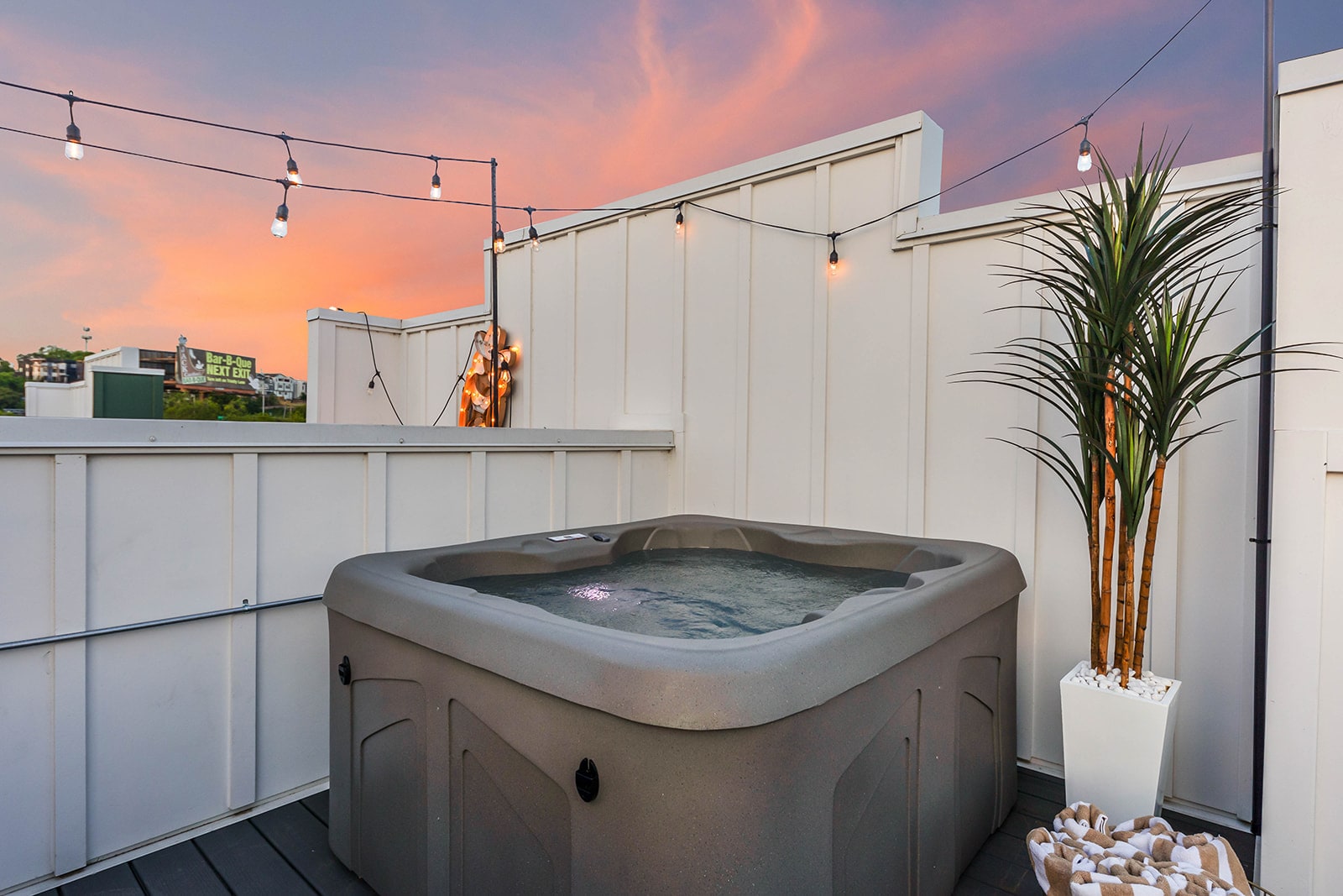 Rooftop hot tub area with a grey hot tub, a tall potted plant, and string lights, set against a beautiful sunset sky with soft pink and orange hues.