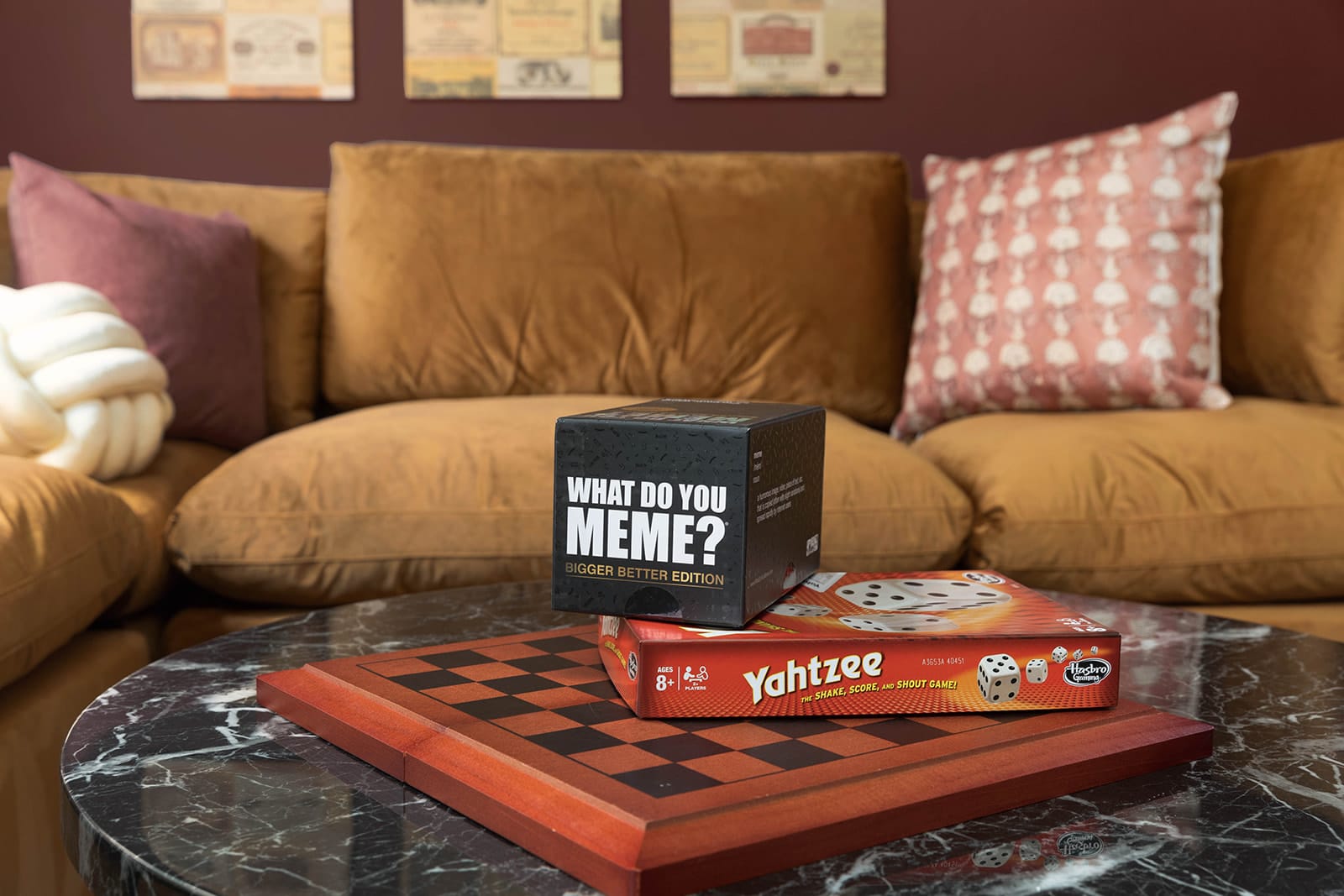 A cozy living room features a large brown corduroy sofa, a marble-top coffee table, and board games (What Do You Meme?, Yahtzee) on a wooden chess board. The space is warmly lit with decorative pillows adding a touch of comfort.
