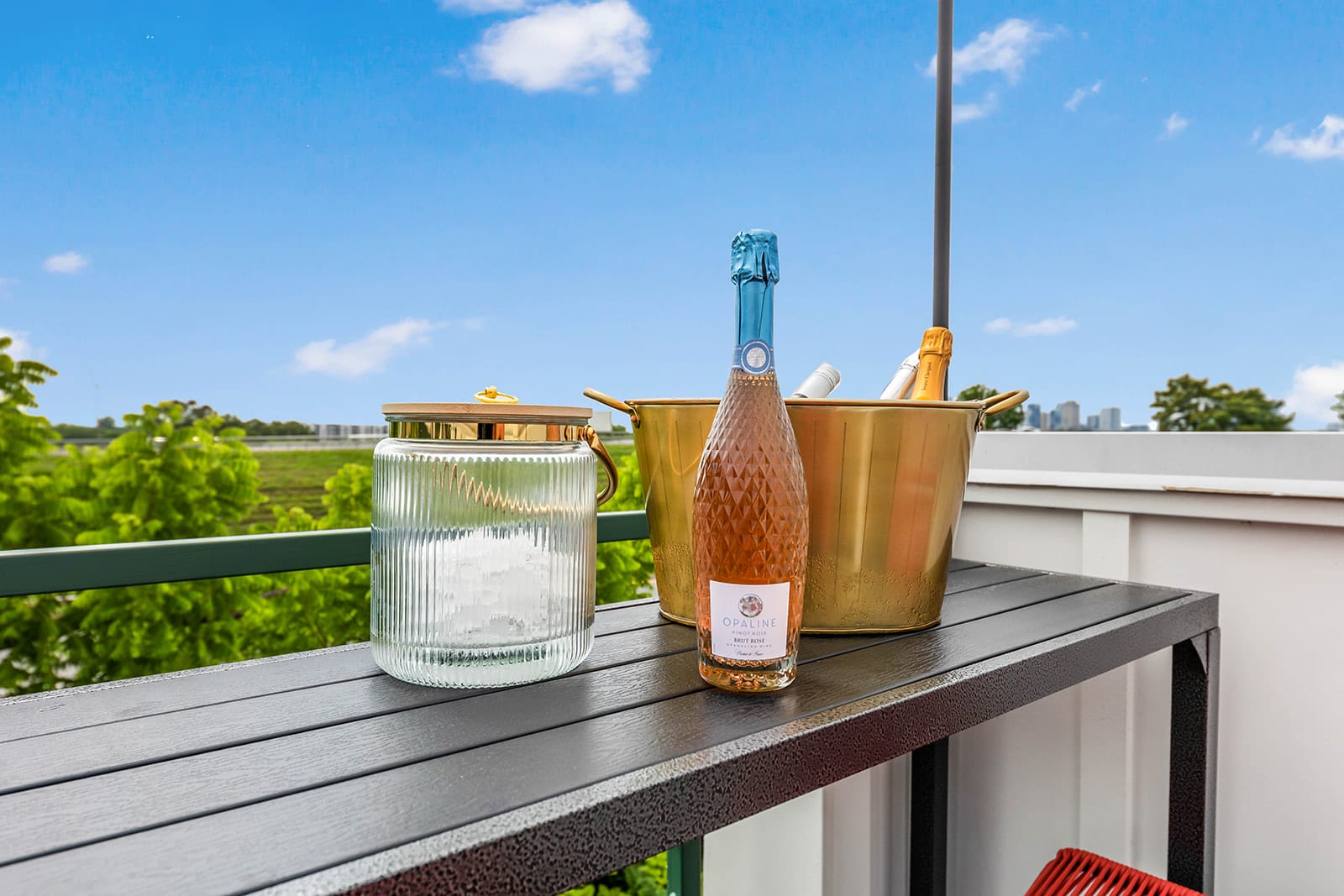 A close-up of a dark slatted outdoor table on a rooftop deck, featuring a bottle of rosé champagne in a gold ice bucket and a clear glass ice bucket, set against a bright blue sky and distant green trees.