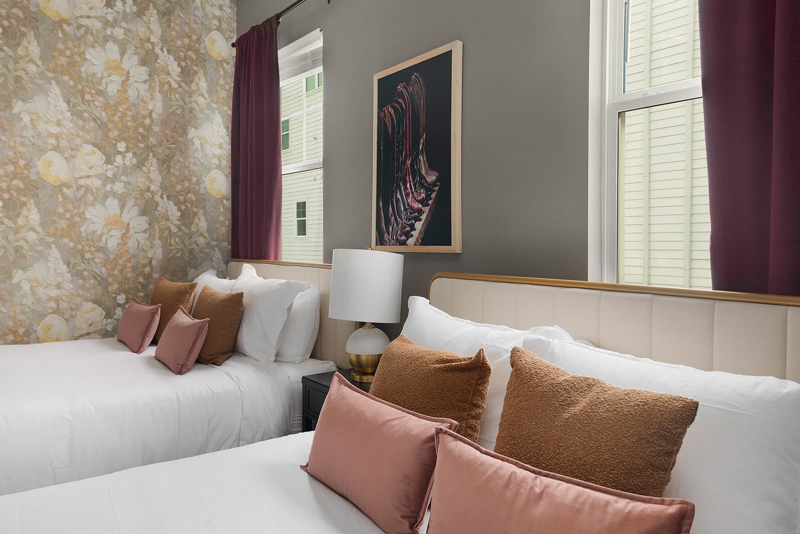 A close-up view of two queen beds in a bedroom, adorned with white bedding, brown textured pillows, and pink decorative pillows. The beds have cream upholstered headboards and gold frames, with a gray wall and burgundy curtains in the background.