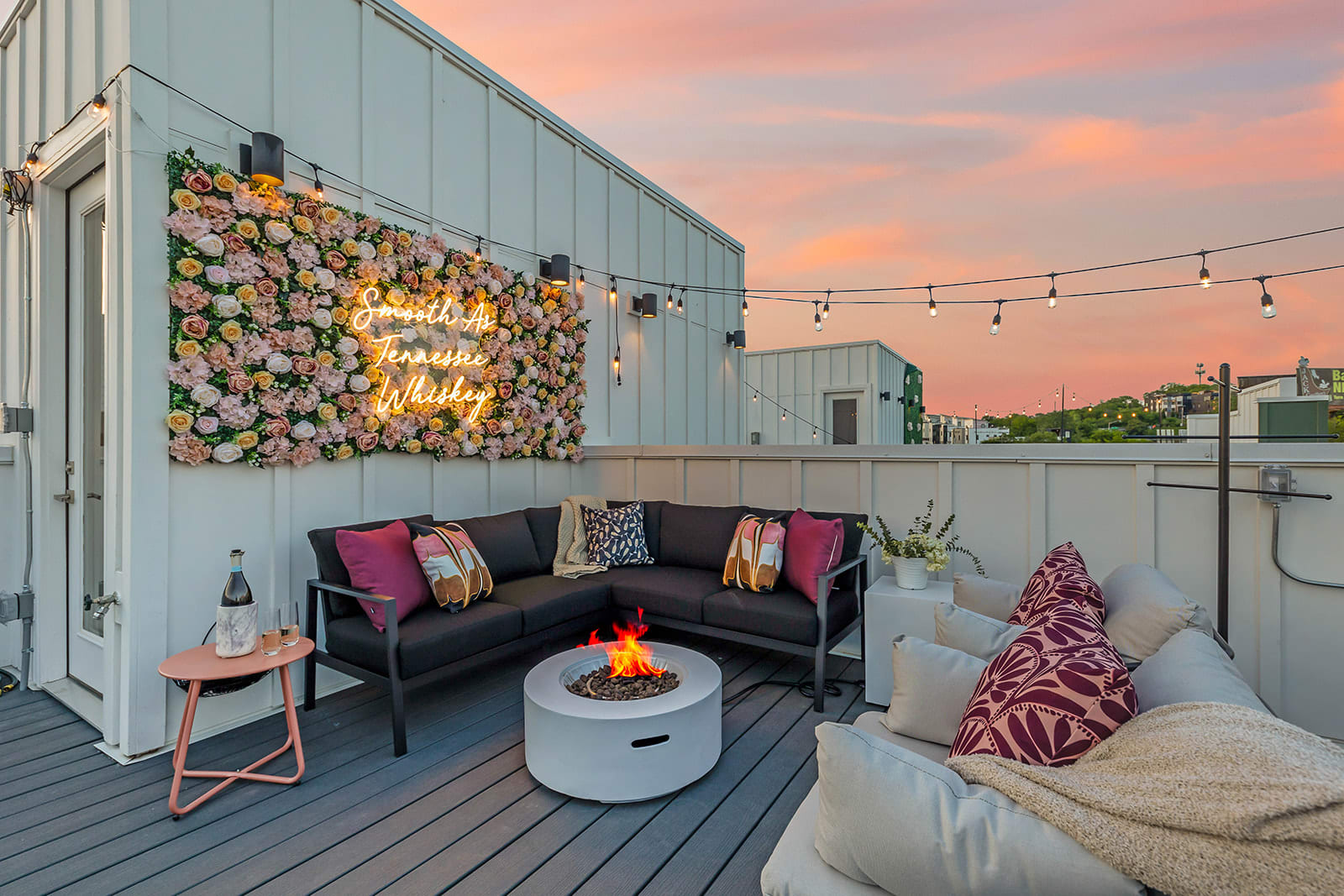 A cozy rooftop deck lounge area features a black sectional sofa, white armchairs, and a circular fire pit, all set against a white wall adorned with a floral arrangement and a neon sign under a sunset sky.