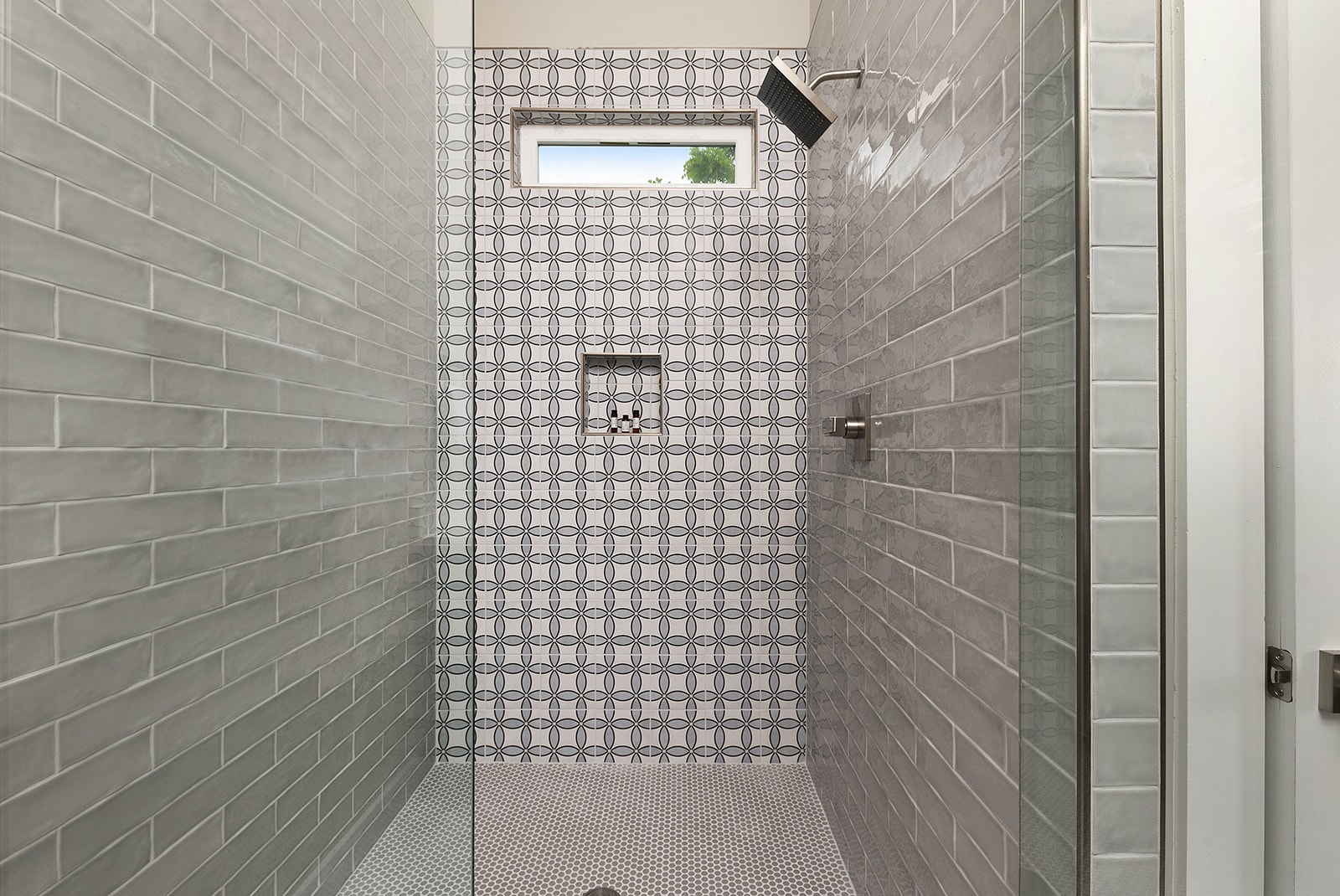 A spacious walk-in shower featuring light gray subway tiles on the walls and a patterned geometric tile accent wall. A small window provides natural light, and a black rain shower head is installed.