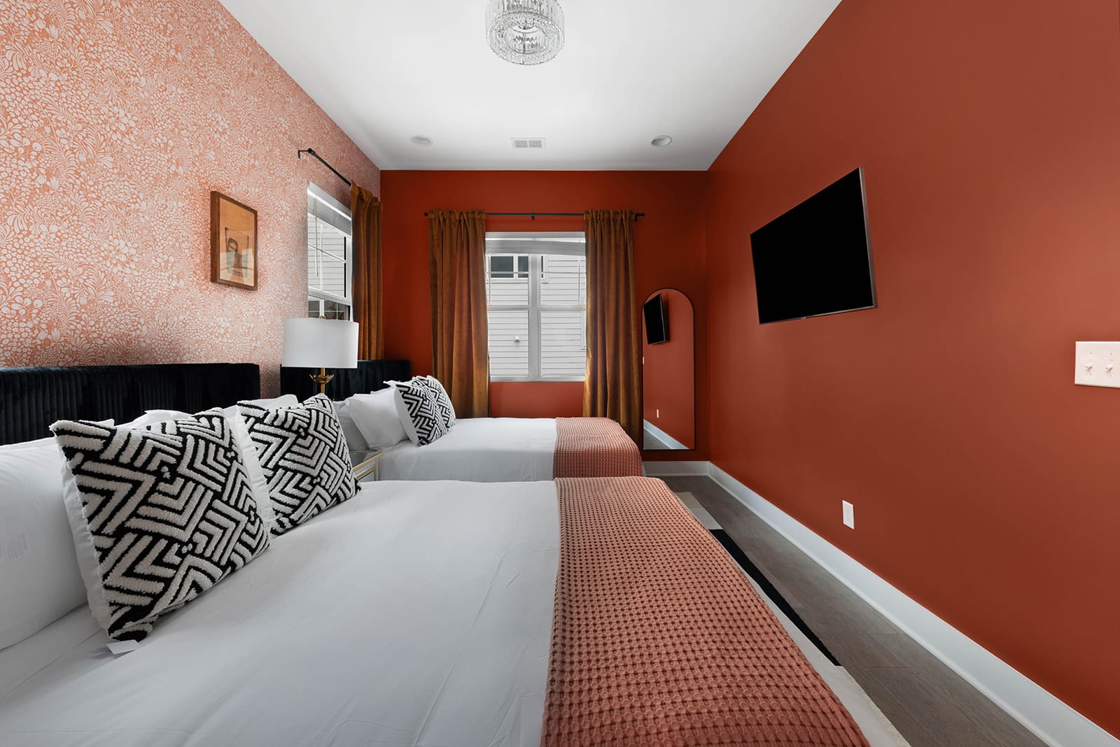 A view of a bedroom with two beds, showcasing a patterned accent wall and solid orange-red walls, complemented by a wall-mounted TV. Each bed is styled with black and white patterned pillows and a soft orange throw.