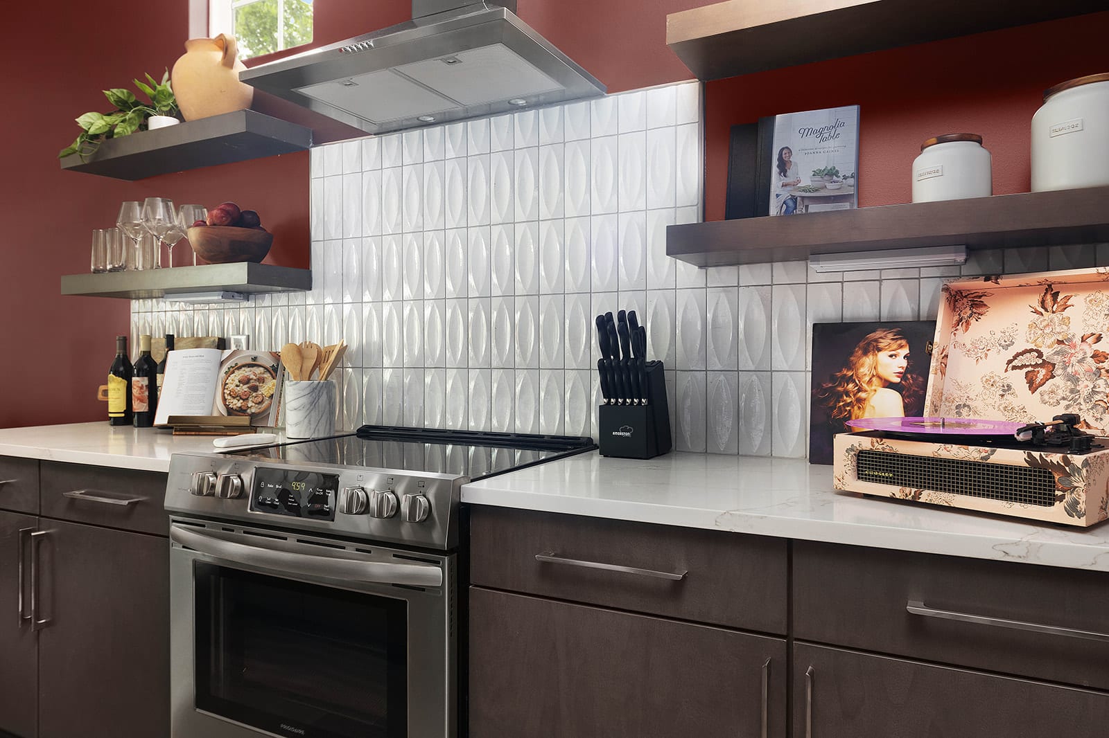 A detailed view of a modern kitchen highlights dark lower cabinets, white quartz countertops, and a textured white tile backsplash. A stainless steel electric range, a knife block, and a vintage-style record player are visible on the counter.