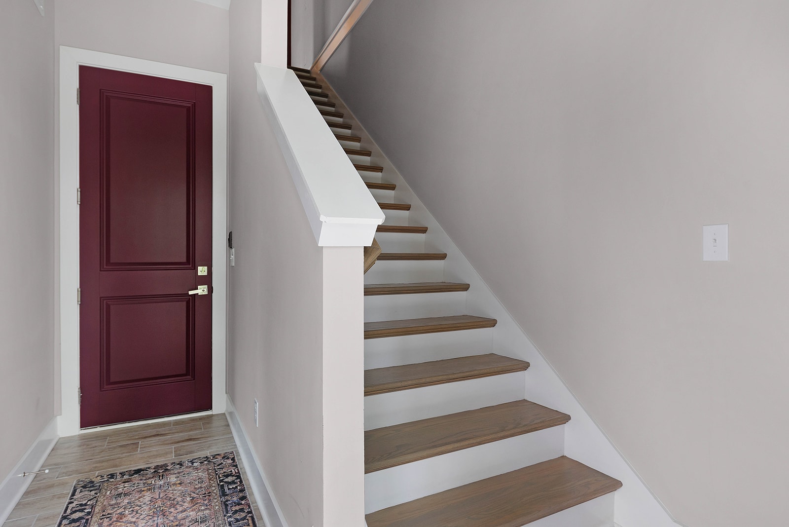A welcoming foyer with a rich burgundy front door, a patterned rug on light wood-look tile flooring, and a staircase with light wood treads and white risers leading upstairs. The walls are painted a light neutral color.