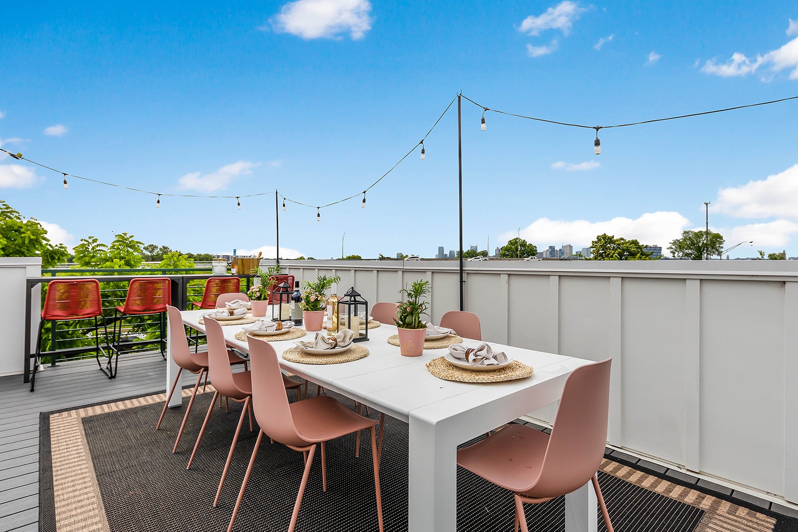 A long white outdoor dining table with pink chairs is set on a dark patterned rug on a rooftop deck, illuminated by string lights overhead and offering distant city views under a bright blue sky.