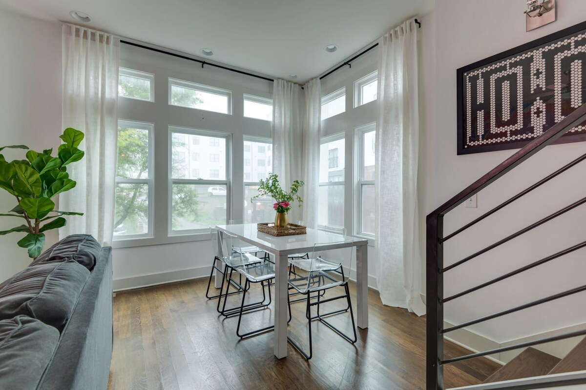 A spacious dining area with a white table and clear chairs is bathed in natural light from numerous large windows with sheer white curtains, offering a glimpse of the living room's dark sofa and a potted plant.