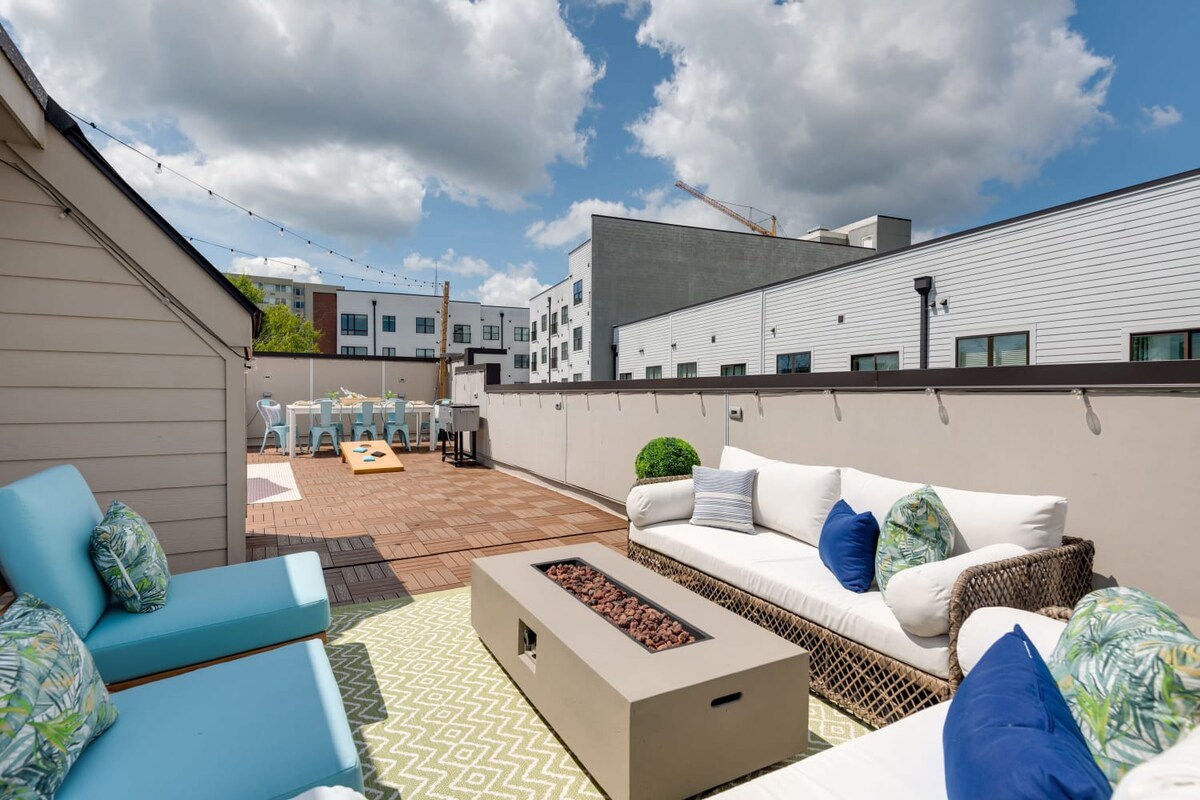 A wide shot of a rooftop deck shows a seating area with a white sofa and blue chairs, a fire pit, and an outdoor dining space with light blue chairs, all under string lights and a bright sky.