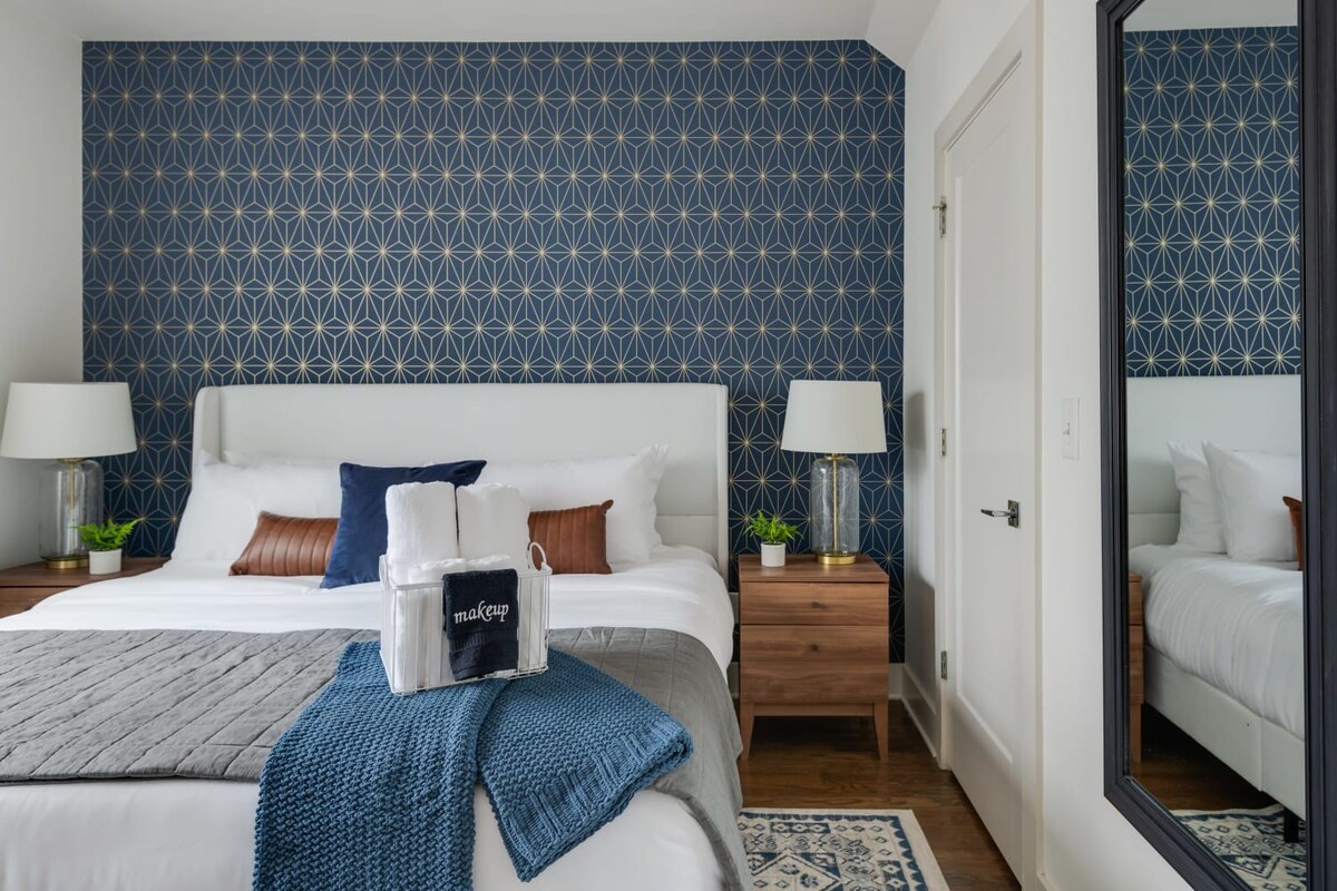 A close-up of the white upholstered bed in a modern bedroom, flanked by wooden nightstands with glass base lamps. The blue and gold geometric accent wall provides a stylish backdrop.