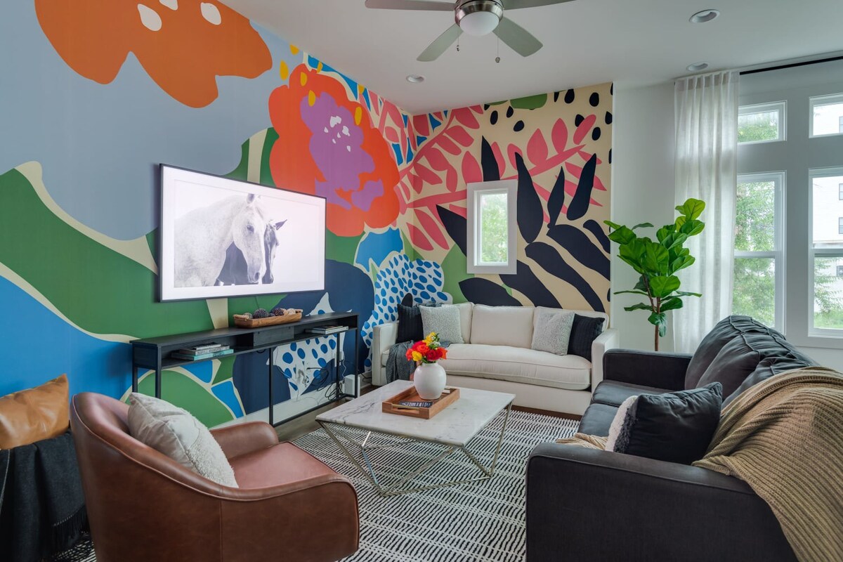 A wide shot of the lively living room showcases a large abstract mural, a white sofa, a dark gray sofa, a brown armchair, and a marble coffee table on a striped rug, with large windows providing natural light.