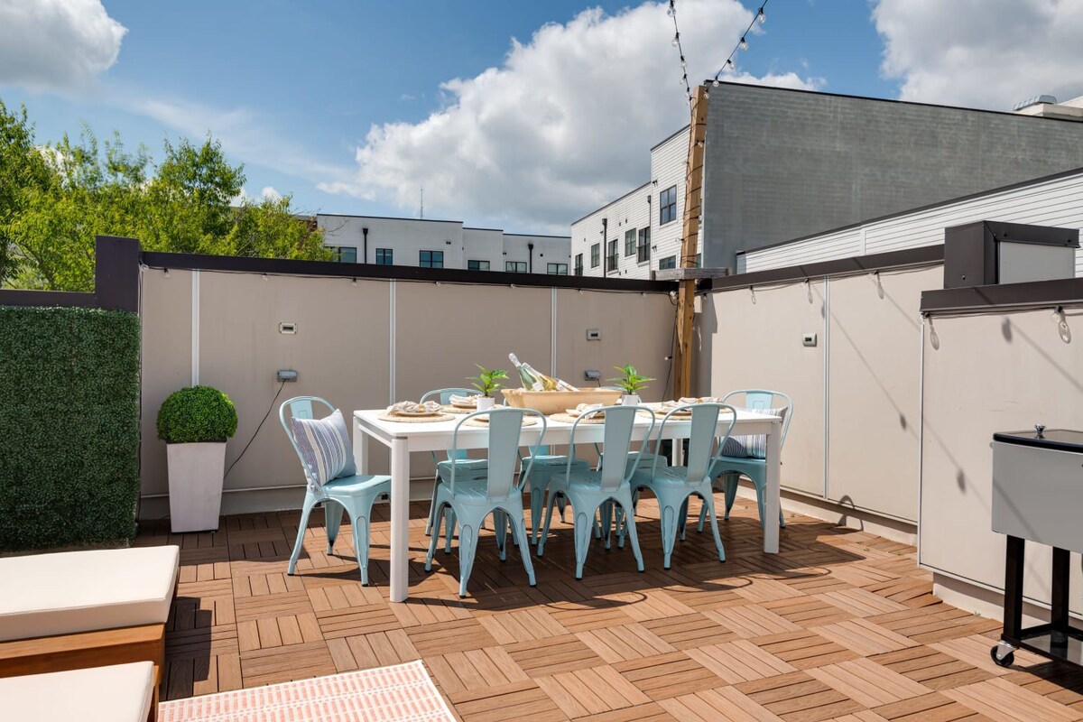 An outdoor dining area on a rooftop deck features a white rectangular table with eight light blue metal chairs, set on a patterned wooden tile floor, with a green faux hedge wall and string lights overhead.