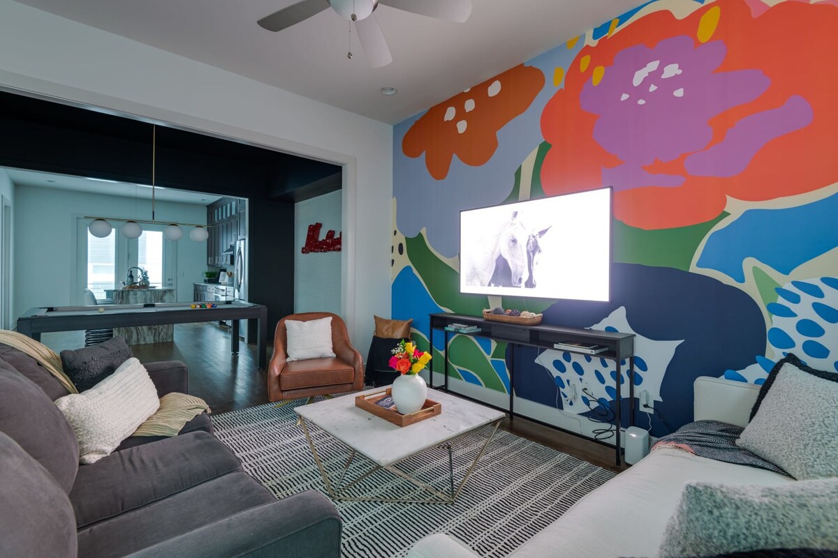 A view of the living room looking back towards the game room, showcasing the open concept layout, the vibrant mural, and the comfortable seating arrangement with a flat-screen TV.