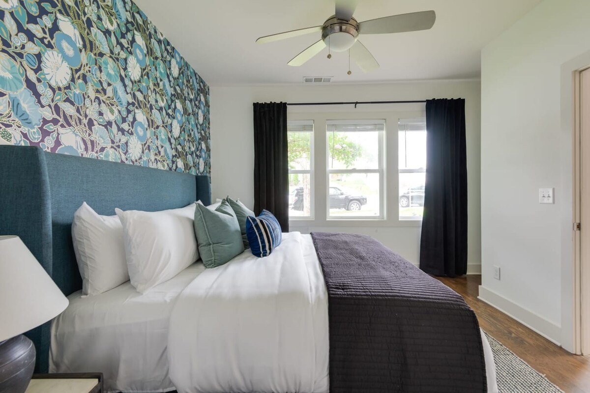 A bright bedroom featuring a large bed with a blue upholstered headboard, white bedding, and a striking blue and green floral accent wall. Black curtains frame a triple window, and a ceiling fan hangs overhead.