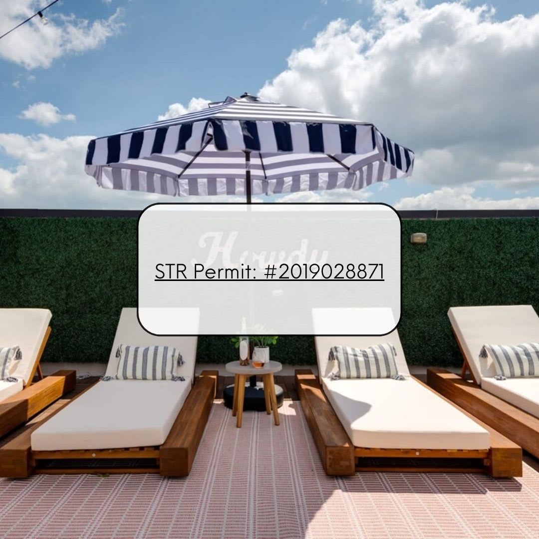 A vibrant rooftop deck features several white lounge chairs with striped pillows, a small side table, and a large black and white striped umbrella, set on a pink and white striped rug. A green hedge wall borders the space.