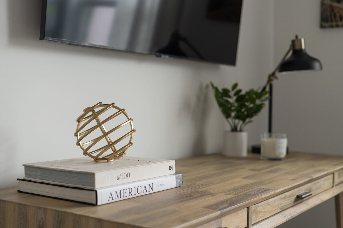 A detailed view of a light wood desk in a bedroom, adorned with decorative books and a gold spherical sculpture. A black task lamp and a small plant add to the functional and aesthetic appeal.