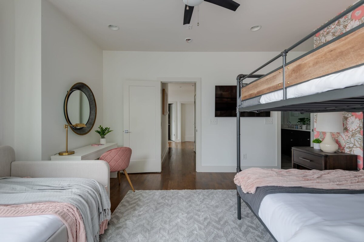 A spacious bedroom designed for multiple guests, featuring a metal bunk bed, a daybed with throw pillows, and a white desk with a round mirror. The room has hardwood floors and a light grey patterned rug.