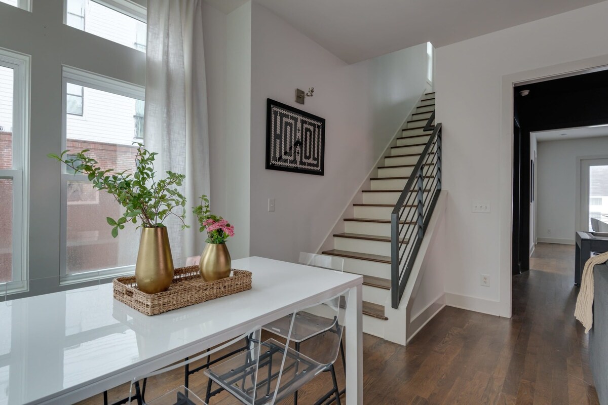 This view of the dining area highlights the white dining table with clear chairs, large windows with sheer curtains, and a staircase with dark wood treads and a black metal railing leading upstairs, with a framed artwork on the wall.