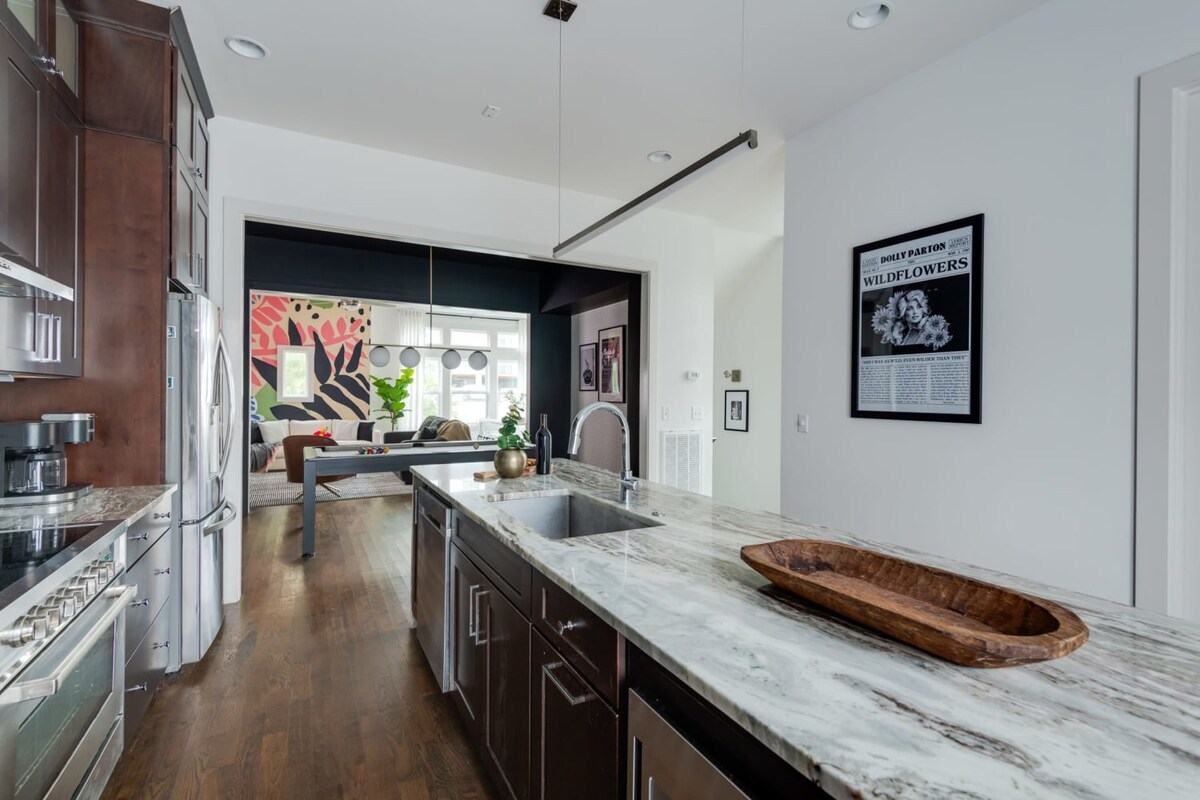A long kitchen island with a marble countertop and sink extends into the foreground, offering a view towards the living room with its vibrant mural and a game room with a pool table, all under a modern linear light fixture.