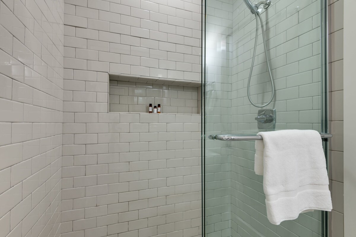 A close-up of a modern glass-enclosed shower with white subway tiles, featuring a built-in niche for toiletries and a handheld showerhead. A white towel hangs on the shower door handle.
