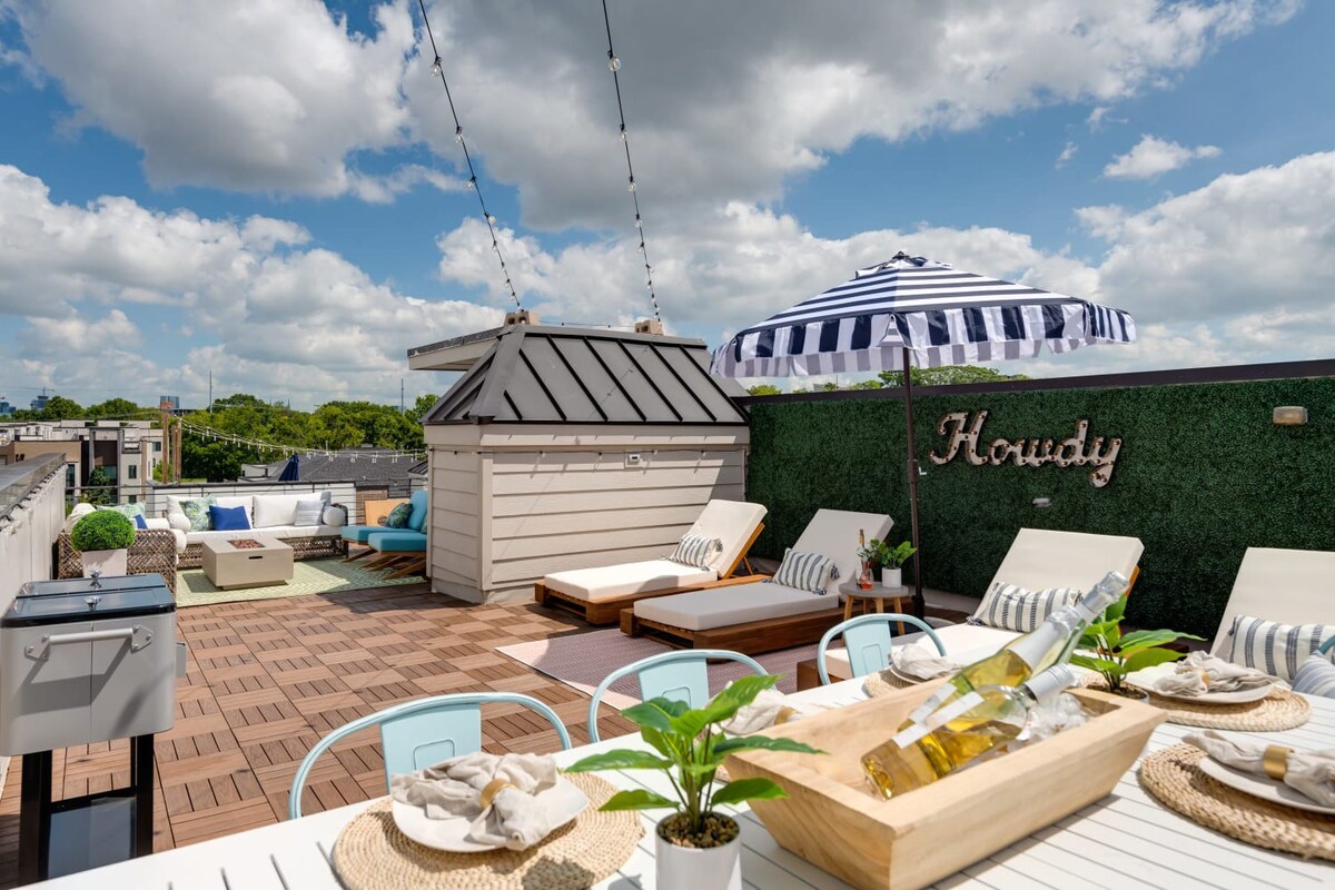 Sunny rooftop deck with a dining area featuring a white table and light blue chairs, lounge chairs, and a striped umbrella, all set on a tiled floor with a green hedge wall.