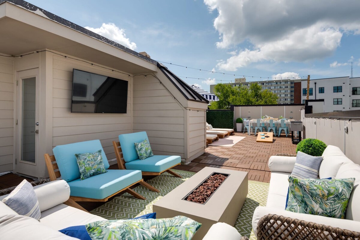 A spacious rooftop deck features a white wicker sofa, light blue lounge chairs, and a fire pit on a green rug, with a wall-mounted TV and string lights overhead, offering a view of an outdoor dining area.