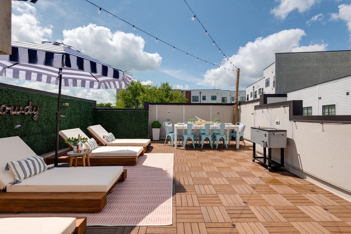 A large rooftop deck features three wooden lounge chairs with white cushions on a pink and white striped rug, a black and white striped umbrella, and an outdoor dining table with light blue chairs.