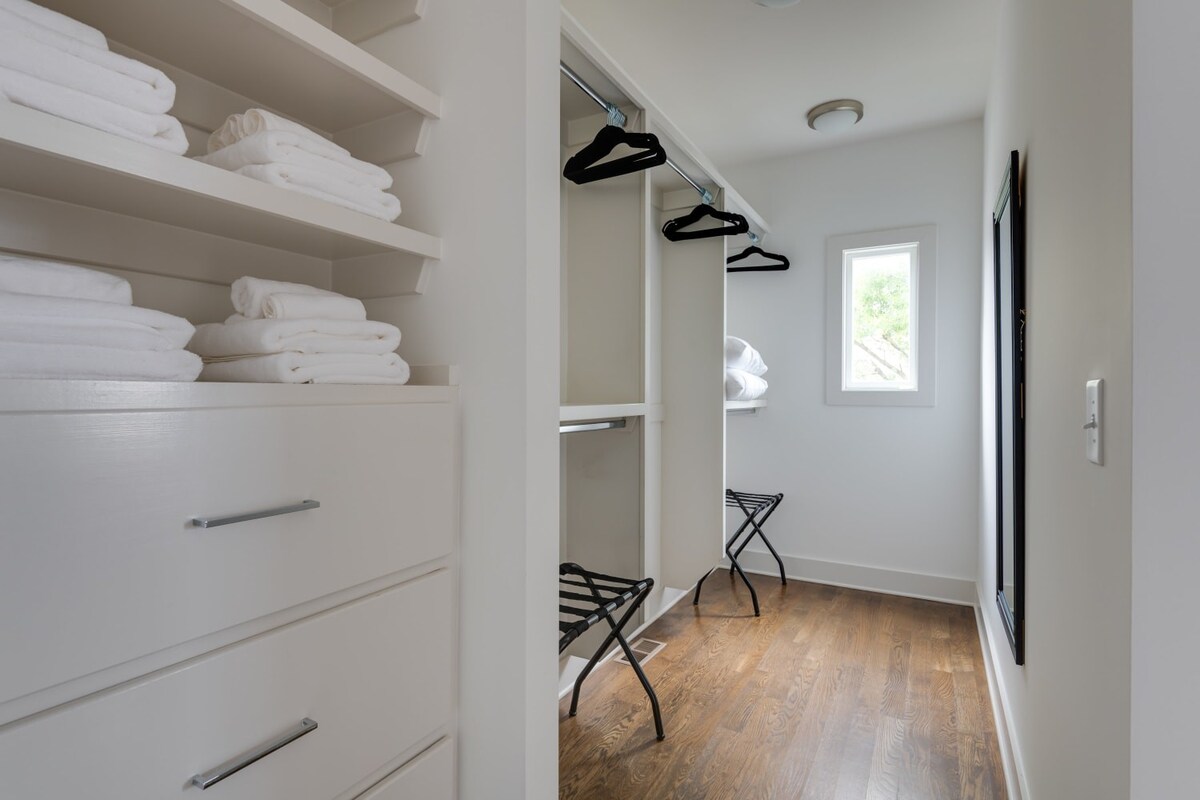 A spacious walk-in closet features white built-in shelving with neatly folded white towels, drawers, and a hanging rod with black hangers. Dark hardwood floors and a small window illuminate the bright space.