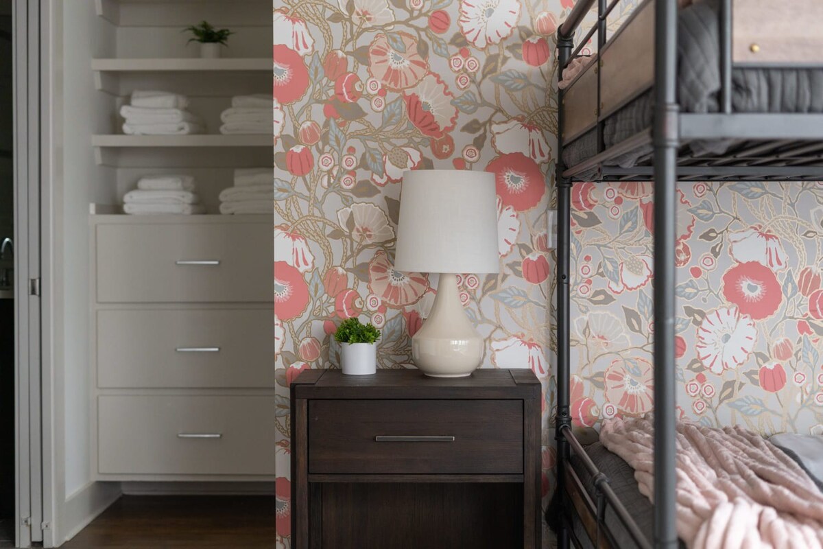 A close-up of a dark wood nightstand with a cream lamp and small plant, set against a vibrant coral floral accent wall. Built-in shelving with neatly folded towels is visible in the background.