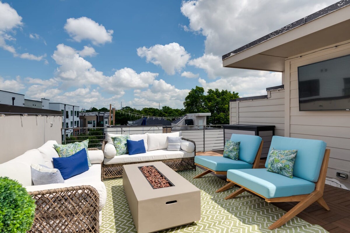 A sunny rooftop deck features a white wicker sofa with blue and green pillows, two light blue lounge chairs, and a rectangular fire pit on a green and white geometric rug, with modern buildings in the background.