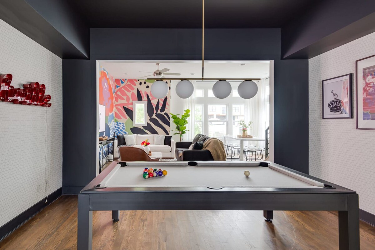 A stylish game room featuring a modern black pool table with a light grey felt, set on dark hardwood floors, with a vibrant, colorful mural visible through a wide archway leading to the living area.