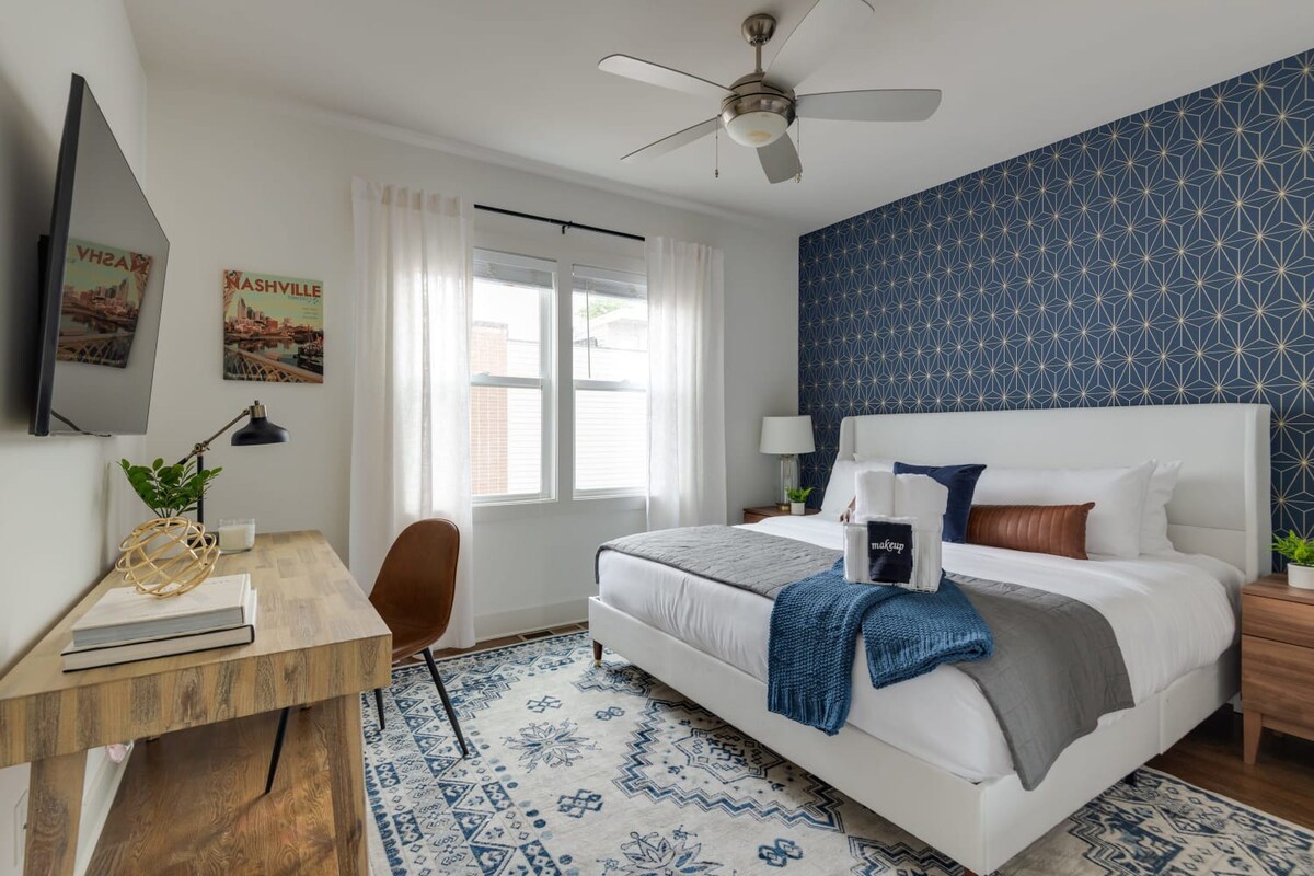 A modern bedroom with a white upholstered bed, blue and gold geometric accent wall, and a light wood desk with a mounted TV. A blue and white patterned rug covers the dark hardwood floor.