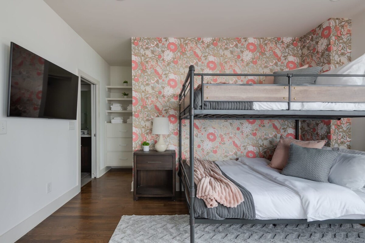 A cozy bedroom featuring a metal bunk bed with wooden accents and a vibrant coral and green floral accent wall. A dark wood nightstand with a lamp sits beside the bed, with built-in shelving visible in the background.