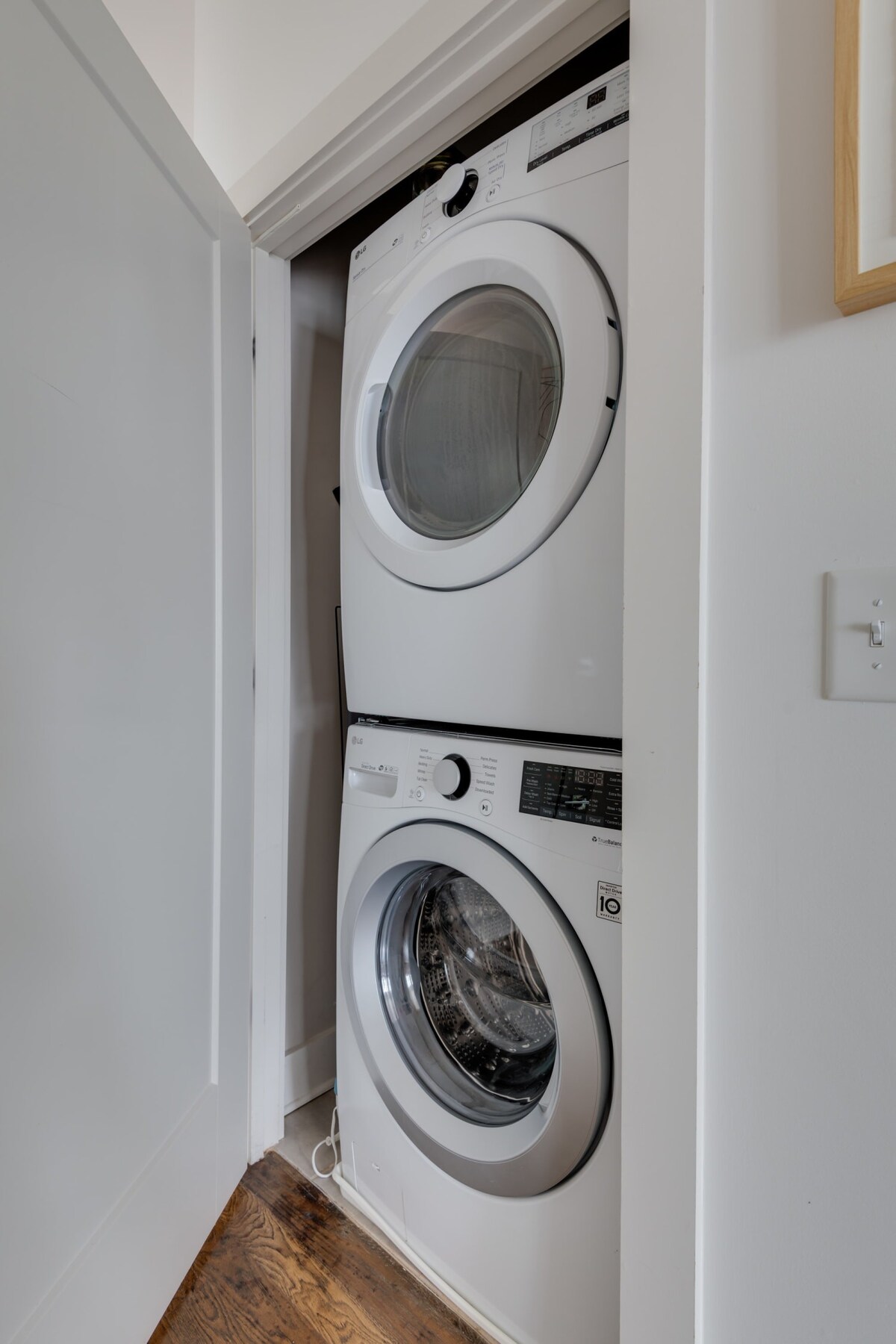 A white stacked washer and dryer unit is neatly tucked into a narrow closet with a light wood floor, seen through an open white door. The modern appliances feature digital displays and front-loading doors.