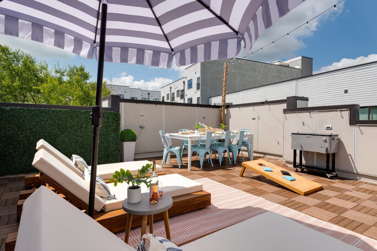 A sunny rooftop deck showcases three wooden lounge chairs with white cushions, a black and white striped umbrella, an outdoor dining table with light blue chairs, and a cornhole game, all on a patterned wooden tile floor.
