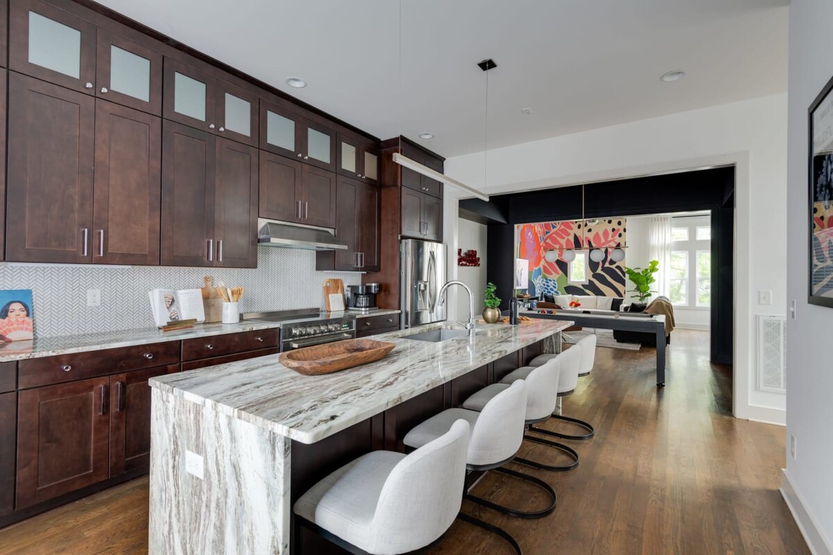 A modern kitchen showcases dark wood cabinetry, white subway tile backsplash, stainless steel appliances, and a large marble-patterned island with four light gray bar stools, opening to a living area with colorful art.
