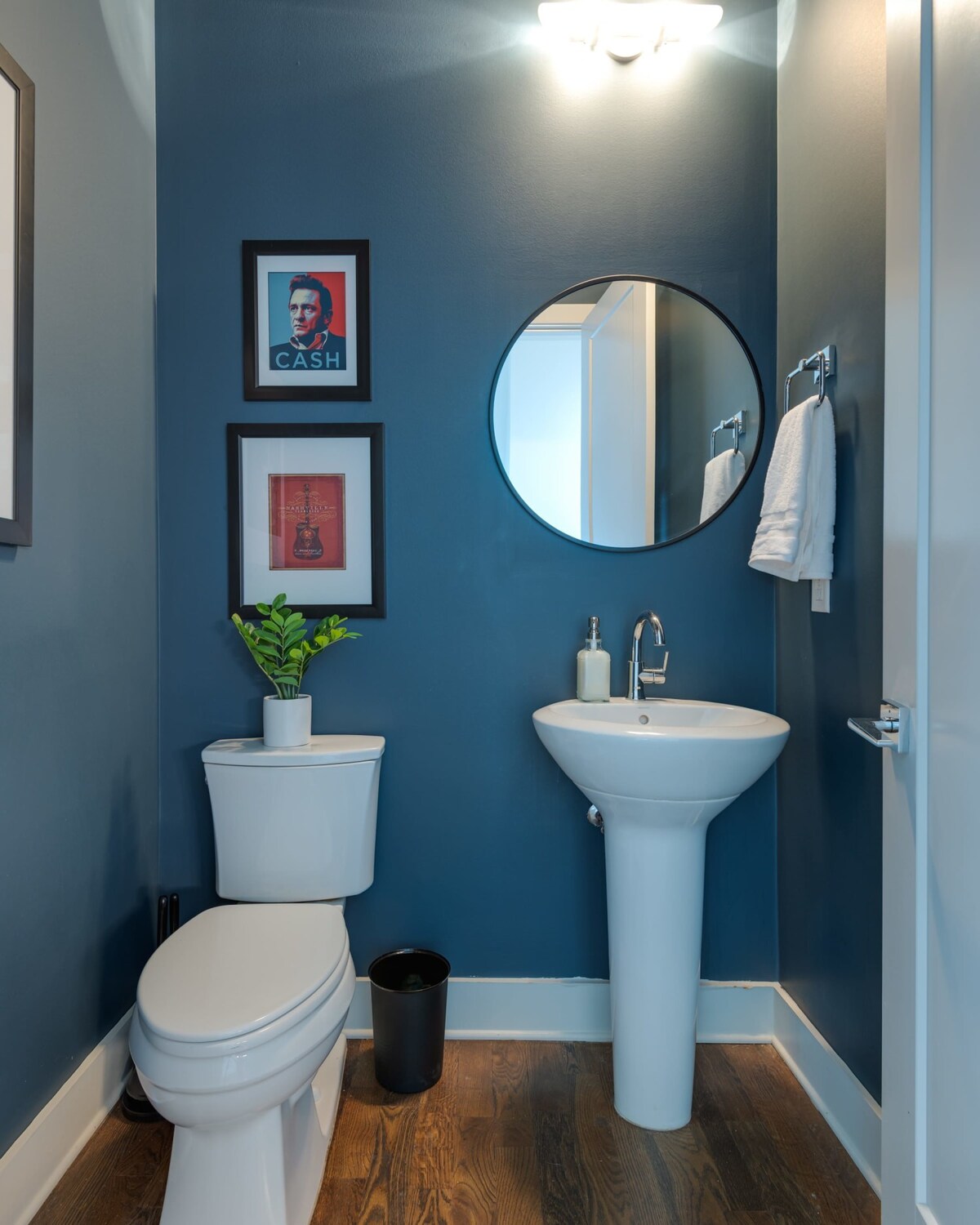 A compact powder room with dark blue walls, a white pedestal sink, a toilet, and a round black-framed mirror. Framed artwork, including a Johnny Cash portrait, decorates the wall.