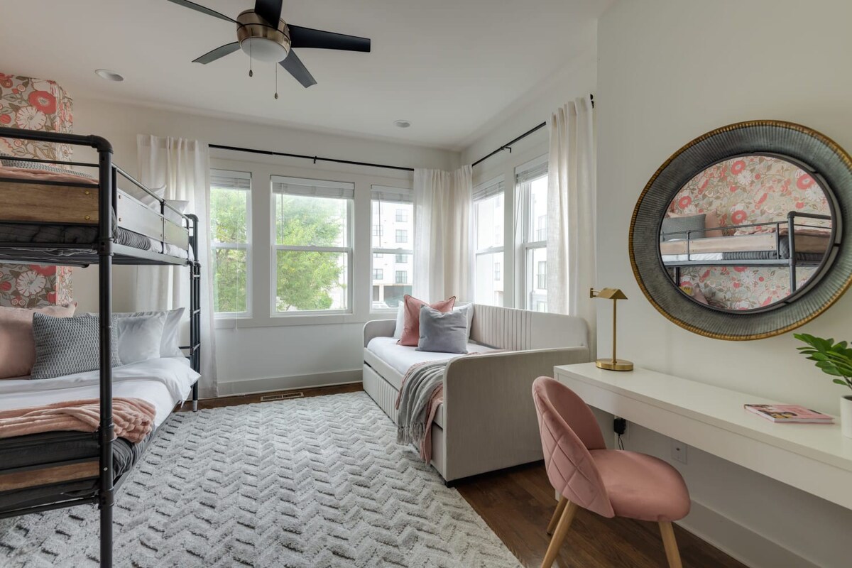 A wide view of a cheerful bedroom featuring a metal bunk bed and a daybed, both dressed with soft pink and grey linens. The room is well-lit by multiple windows and includes a white desk with a round mirror.