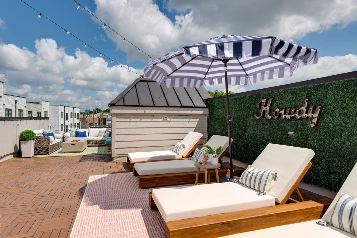 A spacious rooftop deck features three wooden lounge chairs with white cushions on a pink and white striped rug, a black and white striped umbrella, and a "Howdy" sign on a green wall, with a seating area in the background.