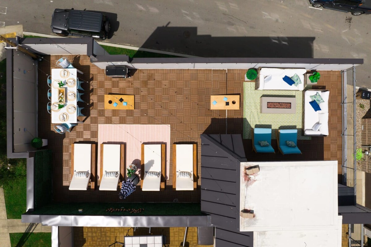An overhead aerial shot directly down onto the rooftop deck, revealing a well-appointed outdoor space with a dining area, lounge seating with a fire pit, sun loungers, and a grill, all on a patterned tile surface.