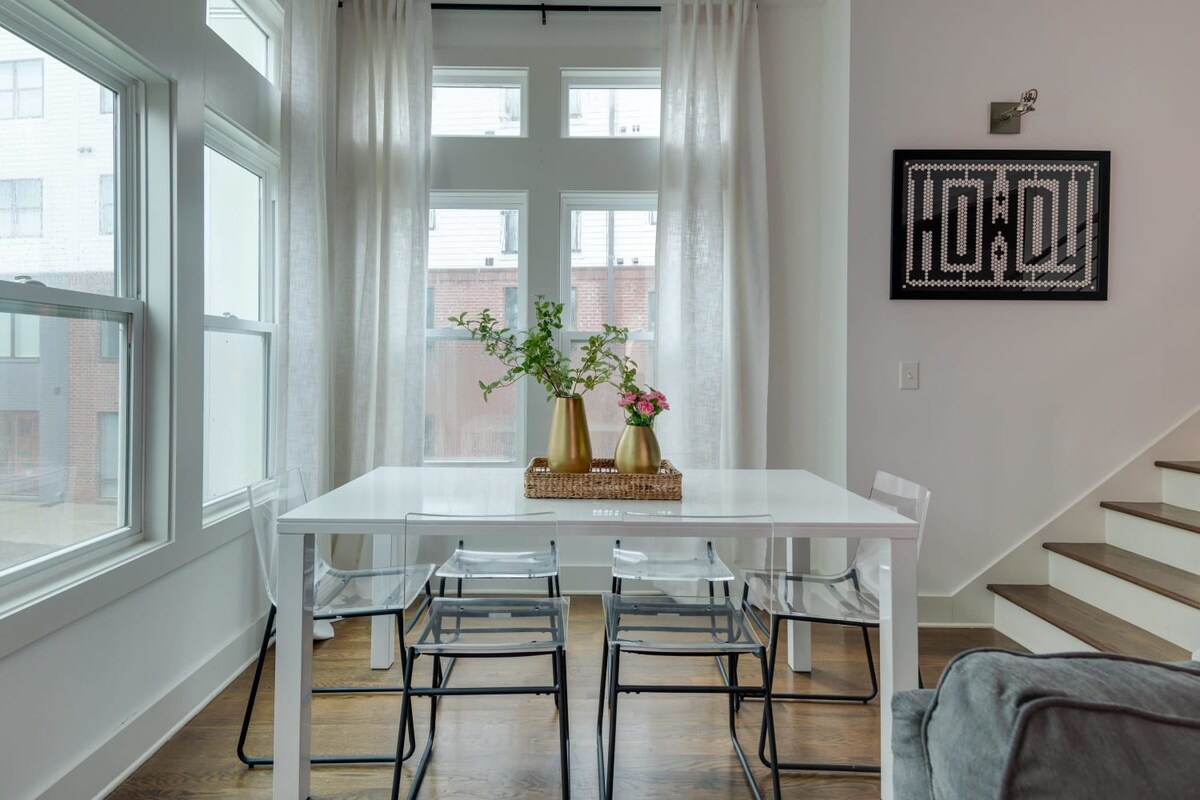 A bright dining area features a white rectangular table with clear acrylic chairs, positioned in a corner with multiple large windows draped with sheer white curtains, and a black framed artwork on the adjacent wall.