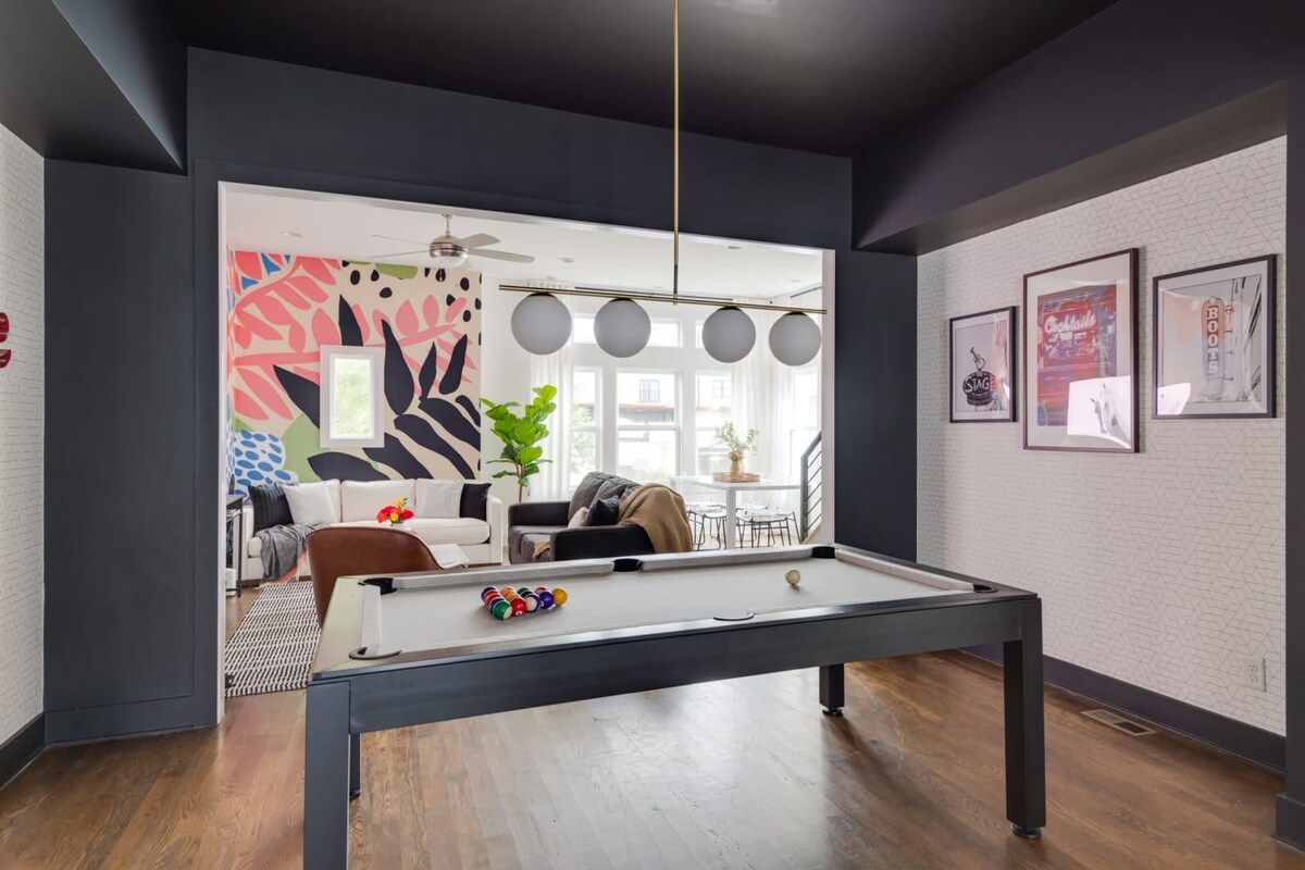 A wide shot of the game room with the pool table, showing the open archway leading to the living room with its vibrant mural and large windows, creating a bright and inviting entertainment space.