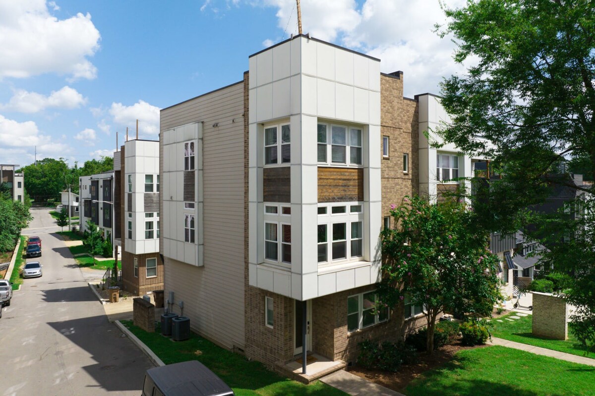 An elevated view captures a row of modern townhouses with varied facades of brick, white siding, and wood, lining a residential street under a partly cloudy sky. Green lawns and mature trees surround the buildings.
