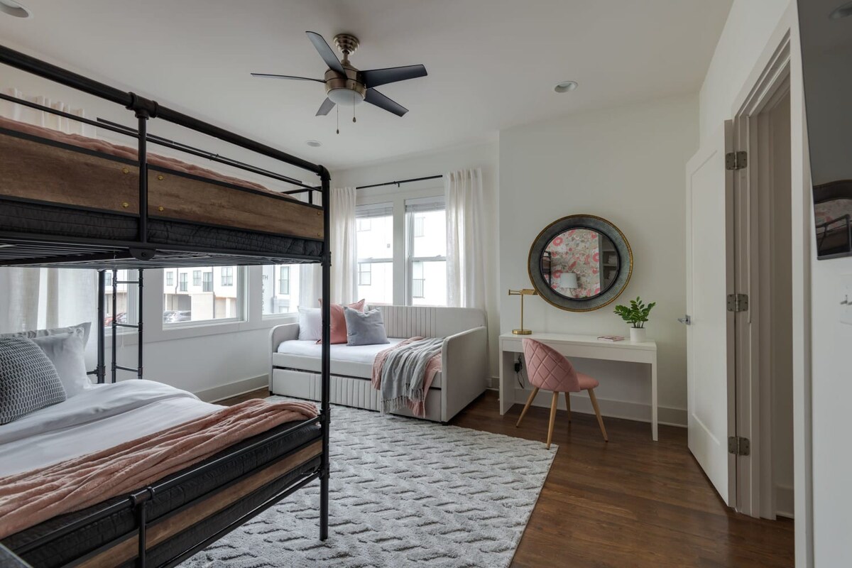 A bright and airy bedroom with a metal bunk bed and a daybed, positioned near large windows with sheer white curtains. A white desk with a pink chair and a round mirror completes the functional and stylish space.