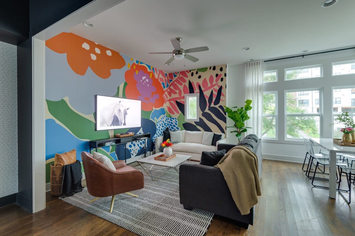 A bright and colorful living room featuring a large abstract floral mural on one wall, a white sofa, a dark grey sofa, and a brown accent chair, all arranged around a marble coffee table on a striped rug.