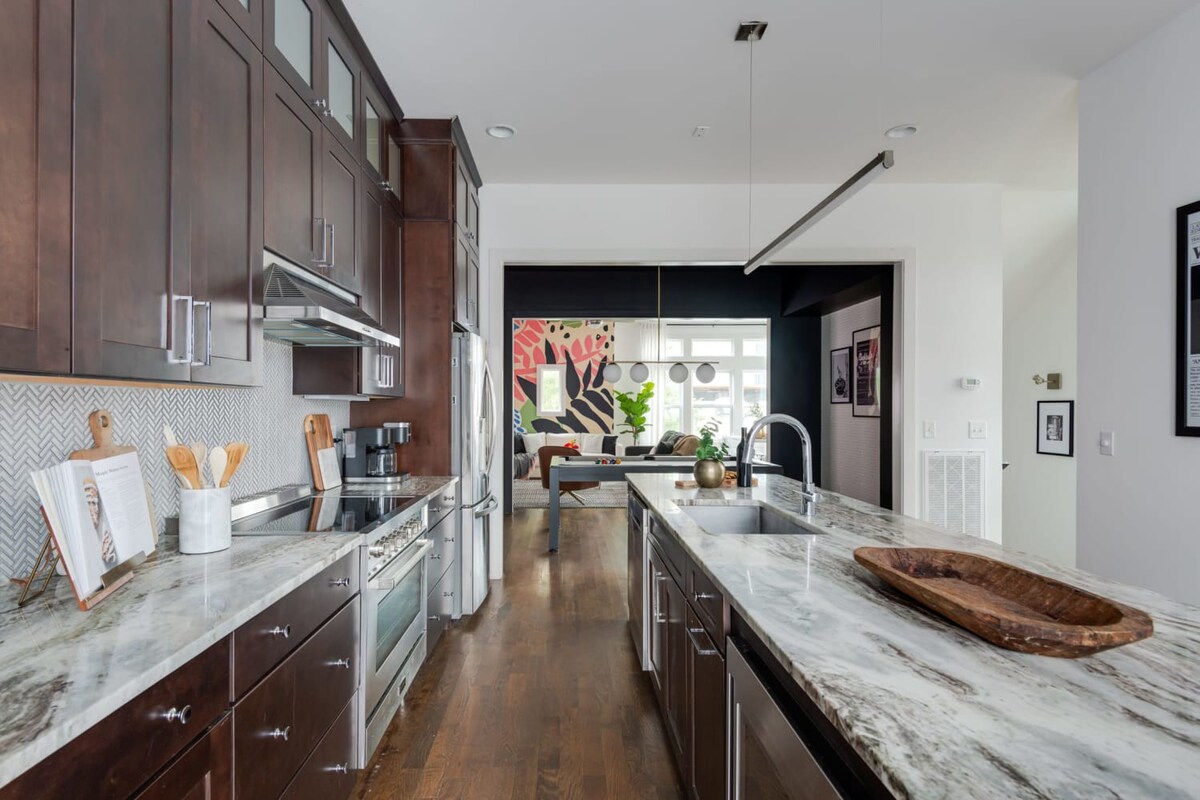 The galley-style kitchen features dark wood cabinets, stainless steel appliances, and a long marble island, providing a clear sightline through an open doorway to the vibrant living room with its colorful mural.
