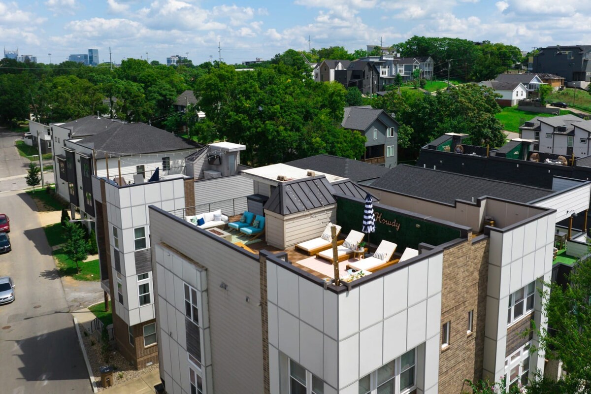 An aerial view of a modern multi-story townhouse with a rooftop deck, featuring outdoor seating and lounge chairs, surrounded by other residential buildings and lush green trees under a partly cloudy sky.