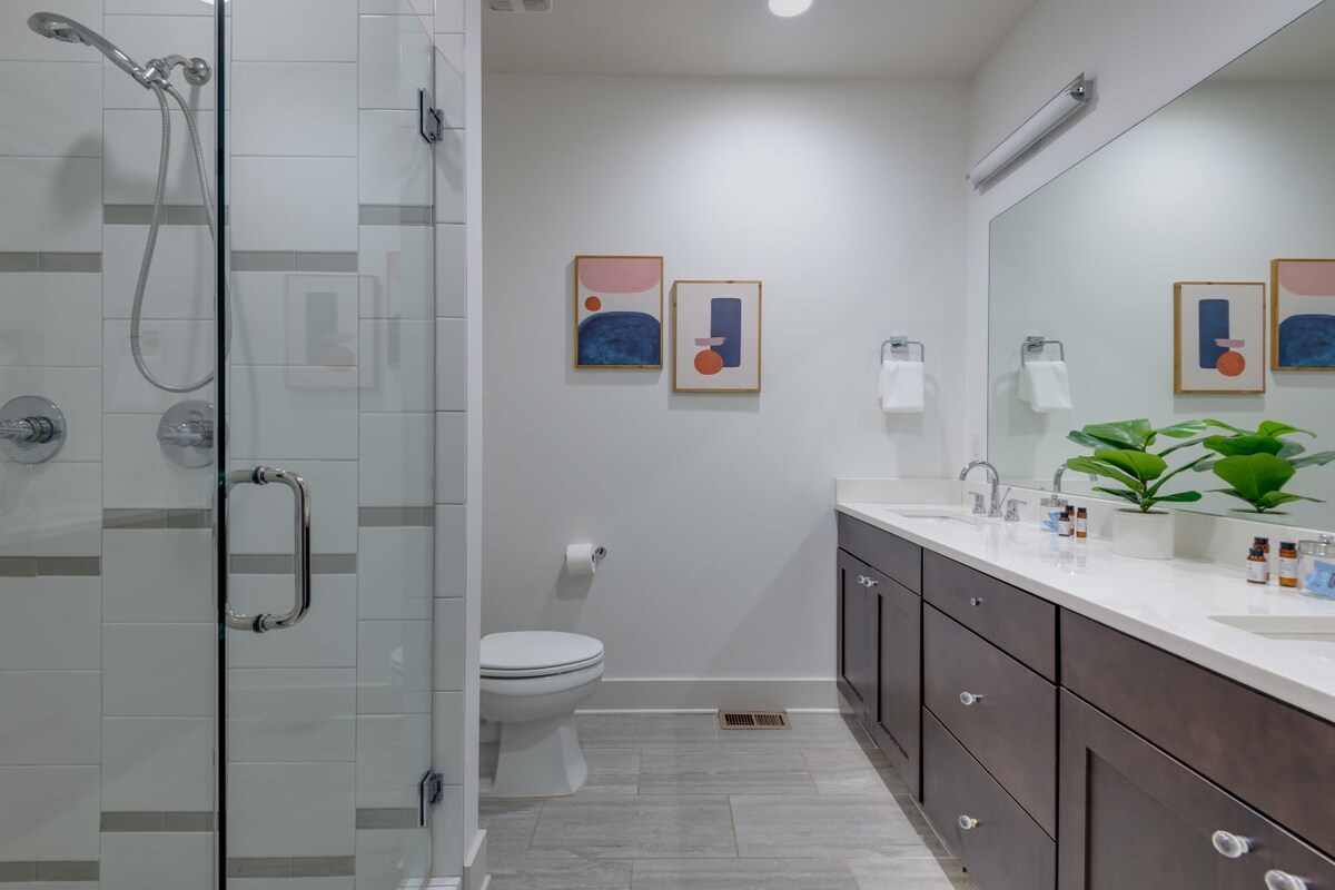 A modern bathroom featuring a glass-enclosed shower with white subway tiles, a white toilet, and a long dark wood double vanity with a white countertop and two sinks. Abstract art pieces adorn the white wall above the toilet.
