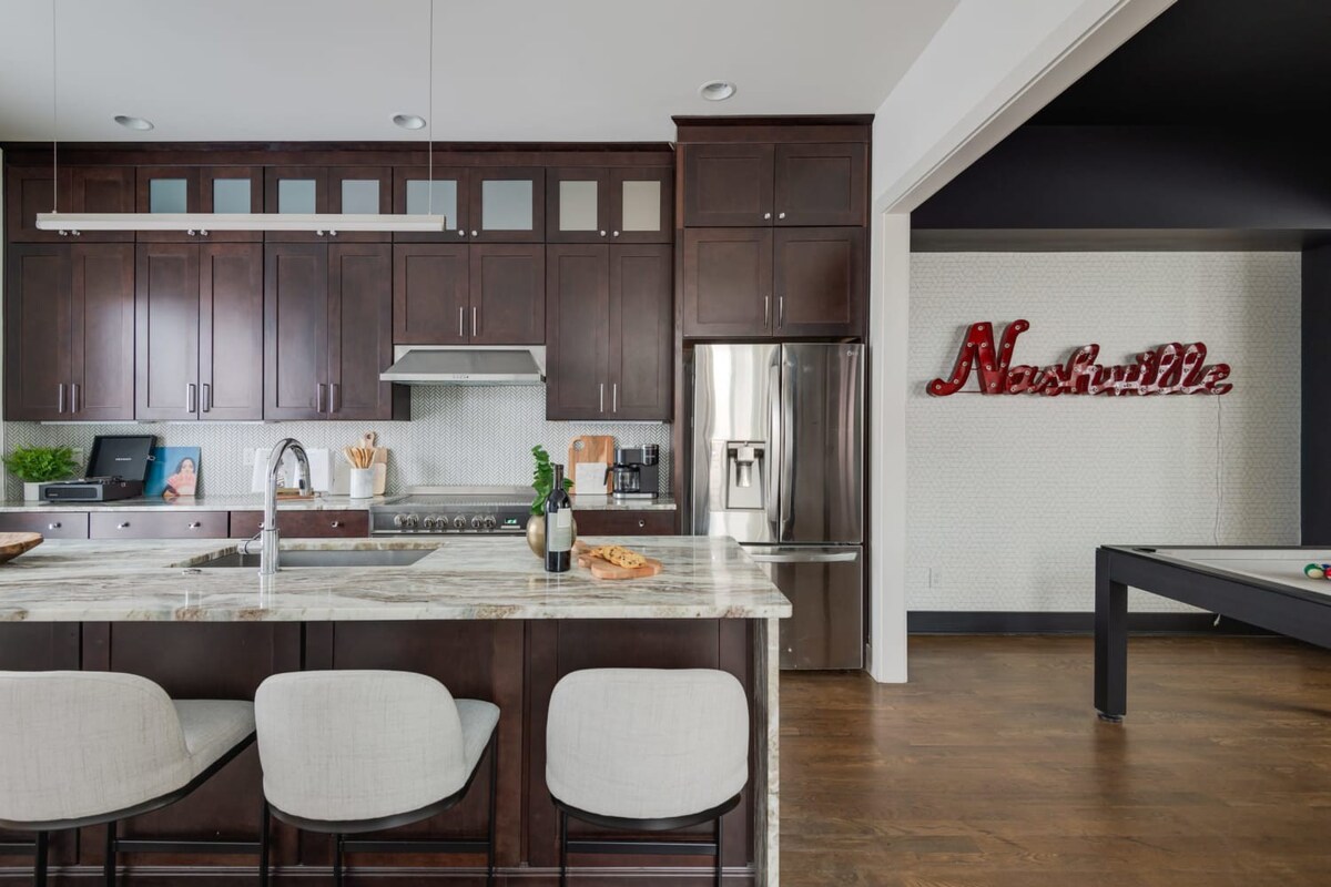 A modern kitchen features dark wood cabinetry, stainless steel appliances including a refrigerator and range, a white hexagonal tile backsplash, and a large island with a light-colored marble countertop and bar stools, opening to a game room.