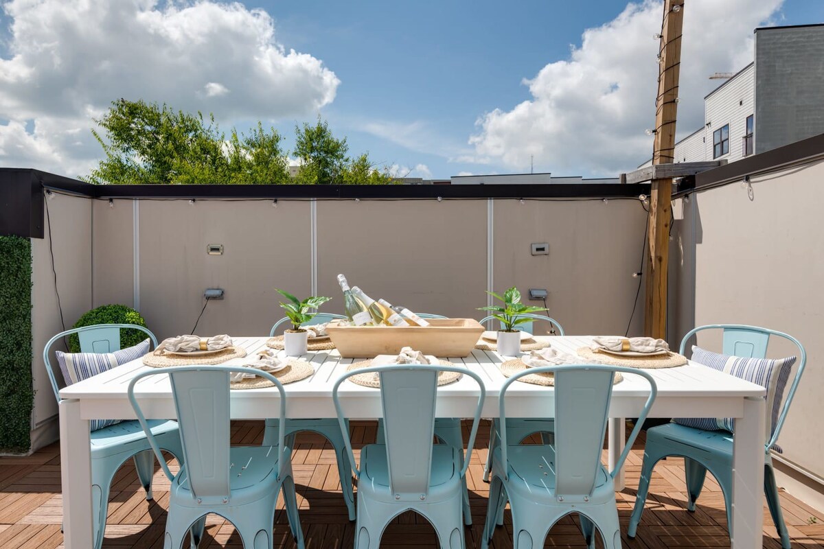 A close-up of an outdoor dining table on a rooftop deck, set with placemats, plates, and a wooden bowl holding wine bottles, surrounded by light blue metal chairs and a green faux hedge wall.