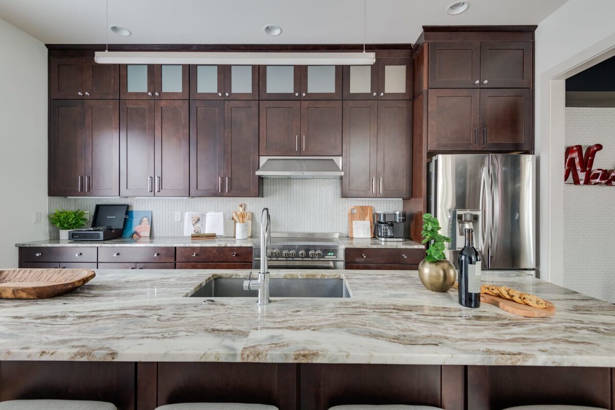 A detailed view of the kitchen shows dark wood cabinets, stainless steel appliances, a white hexagonal tile backsplash, and a large marble island with a sink, adorned with a wooden bowl and a bottle of wine.